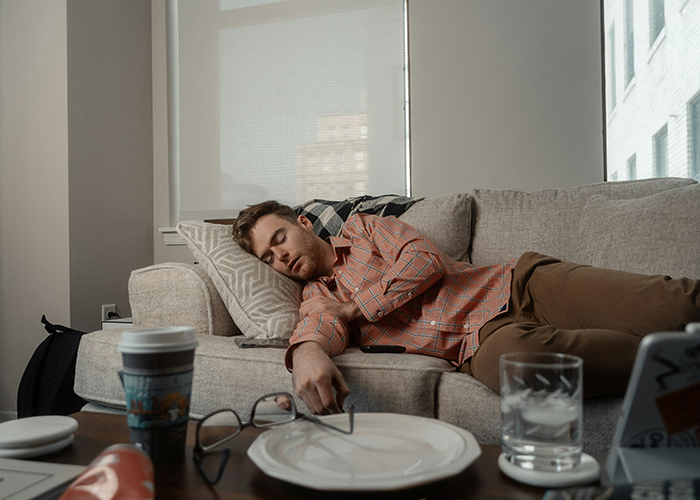 Man sleeping on couch amid household chores, with glasses, coffee, and plate nearby, representing chores and rent themes. Man sleeping on couch amid household chores, with glasses, coffee, and plate nearby, representing chores and rent themes.