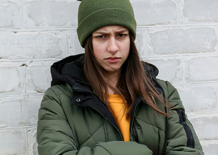 Young woman in a green jacket and beanie, looking upset in front of a brick wall, representing tension over chores and rent. Young woman in a green jacket and beanie, looking upset in front of a brick wall, representing tension over chores and rent.
