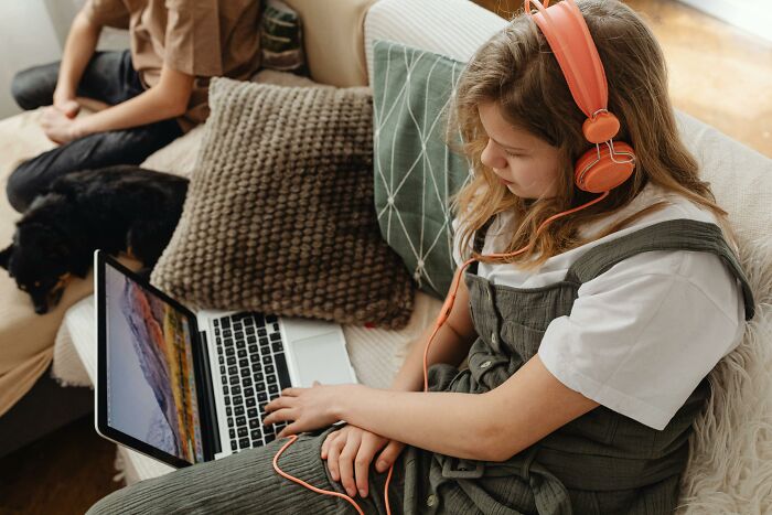 Stepdaughter wearing orange headphones using laptop on couch while stepmom and dog sit nearby during holiday drama. Stepdaughter wearing orange headphones using laptop on couch while stepmom and dog sit nearby during holiday drama.
