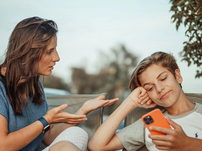 A woman gestures while talking to a teenager with a phone, depicting family dynamics and potential exclusion.