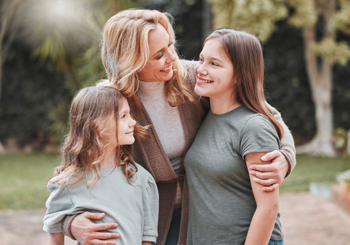 Stepmom outdoors with her kids, smiling and embracing them warmly in a garden setting. Stepmom outdoors with her kids, smiling and embracing them warmly in a garden setting.