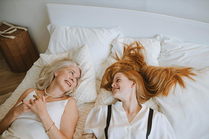 Stepmom and stepdaughter lying on a bed, smiling at each other, surrounded by white pillows. Stepmom and stepdaughter lying on a bed, smiling at each other, surrounded by white pillows.