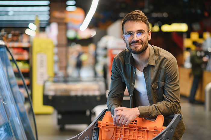 Man in glasses and casual shirt at grocery store with cart, representing man asking a stranger to pay for his groceries.
