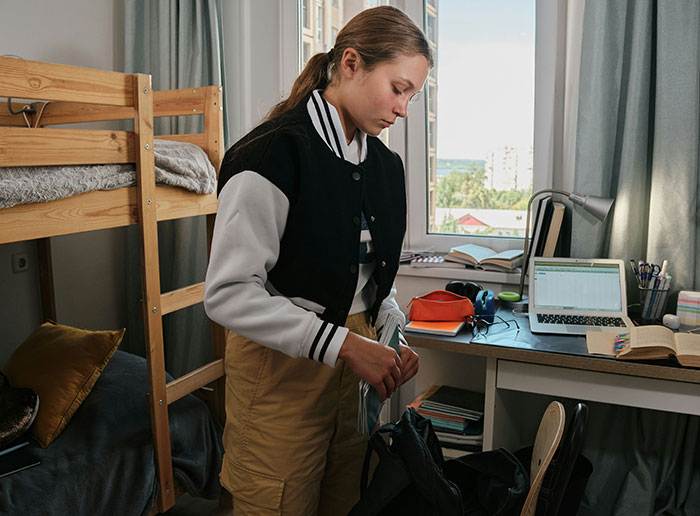 Student organizing items in a college dorm room with a loft bed and study desk visible. Student organizing items in a college dorm room with a loft bed and study desk visible.