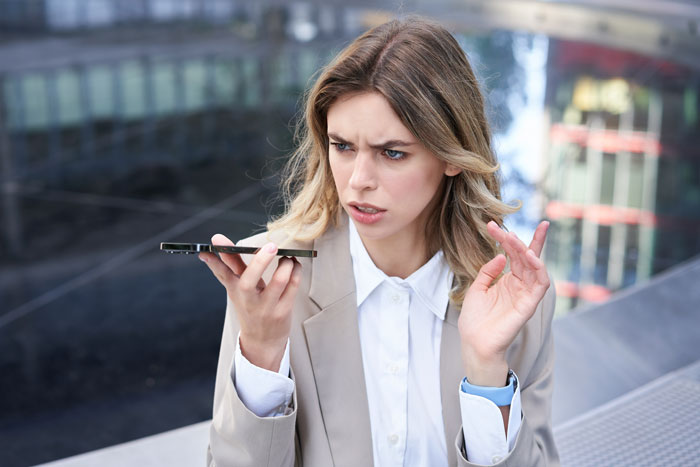 Woman looking at phone with a confused expression, outdoors, wearing a beige blazer, related to engagement photo issue. Woman looking at phone with a confused expression, outdoors, wearing a beige blazer, related to engagement photo issue.