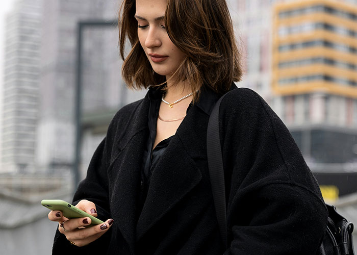 Woman in a black coat looking at her phone, with city buildings in the background, connected to a will and family affairs theme. Woman in a black coat looking at her phone, with city buildings in the background, connected to a will and family affairs theme.