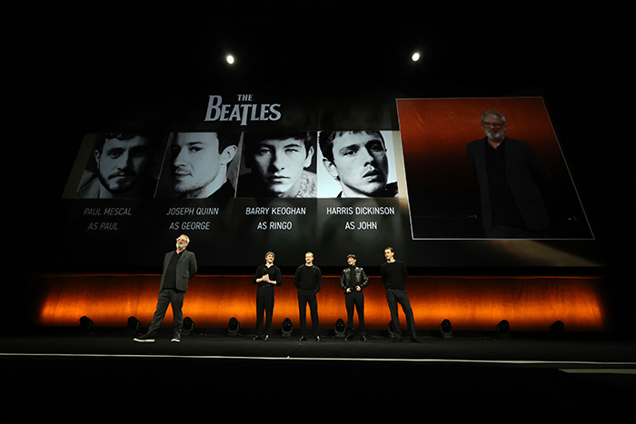 Casting announcement for The Beatles biopic, featuring actors on stage with projected images of their roles. Casting announcement for The Beatles biopic, featuring actors on stage with projected images of their roles.