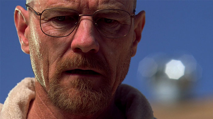 A man with glasses, facial hair, and a towel around his neck, set against a clear blue sky.