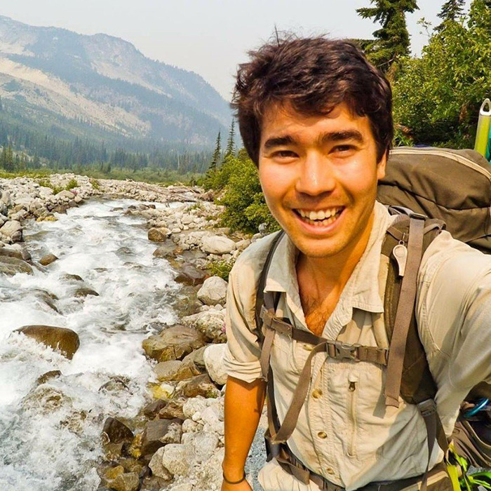 Smiling YouTuber on a mountain trail with river in background, carrying a backpack. Smiling YouTuber on a mountain trail with river in background, carrying a backpack.