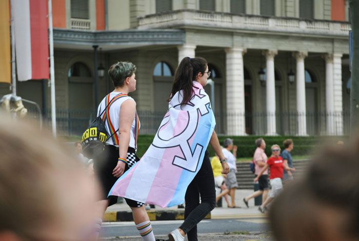 Two people walking, one with trans flag around shoulders, reflecting on trans rights after Supreme Court ruling. Two people walking, one with trans flag around shoulders, reflecting on trans rights after Supreme Court ruling.
