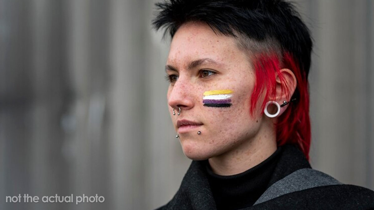 Person with piercings and red-black hair, with a nonbinary pride flag painted on their cheek, showing support for trans topics.