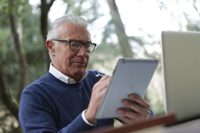 Elderly man with glasses using a tablet outdoors, seated near a laptop. Elderly man with glasses using a tablet outdoors, seated near a laptop.