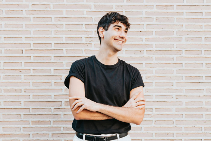 A smiling man in a black shirt stands against a brick wall outdoors, illustrating a story on being gay. A smiling man in a black shirt stands against a brick wall outdoors, illustrating a story on being gay.