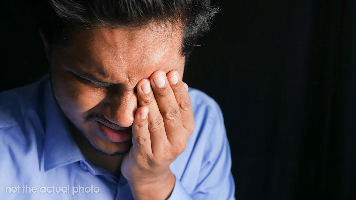 Young man in a blue shirt showing facial expression of deep pain and discomfort against a dark background