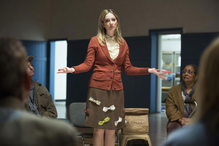 Woman in a classroom setting, wearing a brown and red outfit, gestures with arms outstretched. Woman in a classroom setting, wearing a brown and red outfit, gestures with arms outstretched.