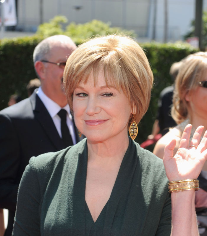 Mary Kay Place smiling outdoors in a green outfit, waving to the camera. Mary Kay Place smiling outdoors in a green outfit, waving to the camera.