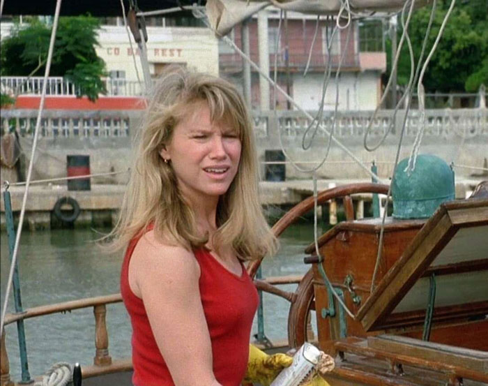 A woman with blonde hair in a red tank top, standing on a boat, wooden deck visible in the background. A woman with blonde hair in a red tank top, standing on a boat, wooden deck visible in the background.