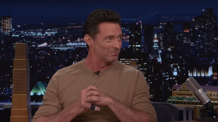 Celebrity in a beige shirt on a talk show set with a cityscape backdrop.