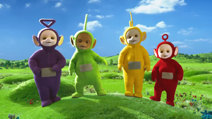 Four colorful Teletubbies standing on a grassy hill under a clear blue sky.