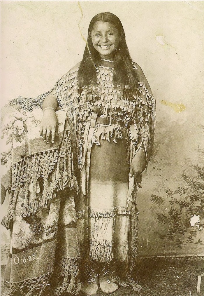 Native American woman in traditional attire, smiling, captured in a haunting photo from a century ago.