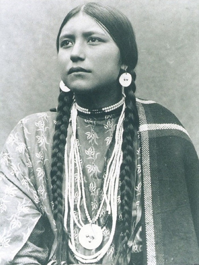 Native American woman from a century ago wearing traditional jewelry and clothing, captured in a haunting historical photo.
