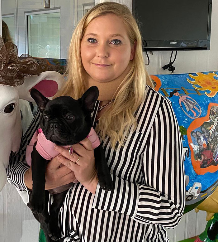 Woman in a striped shirt holding a black dog, next to a decorative cow, related to Prince Andrew's accuser news. Woman in a striped shirt holding a black dog, next to a decorative cow, related to Prince Andrew's accuser news.