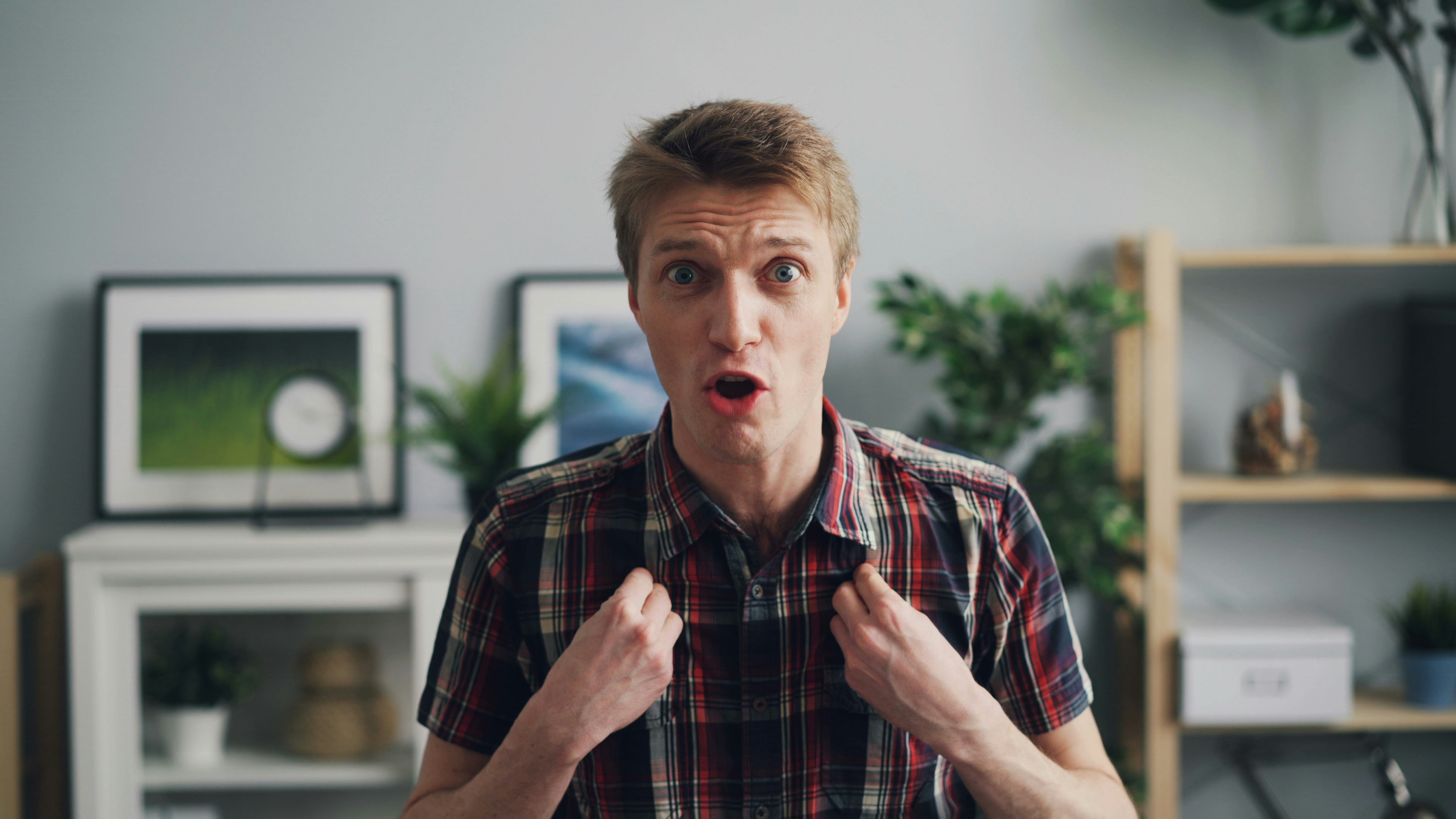 A surprised man in a plaid shirt gestures emotionally, set against a home office background with plants and shelves. A surprised man in a plaid shirt gestures emotionally, set against a home office background with plants and shelves.