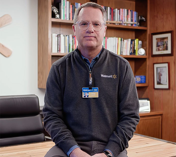Man in a Walmart sweater with an ID badge, seated in an office setting. Man in a Walmart sweater with an ID badge, seated in an office setting.