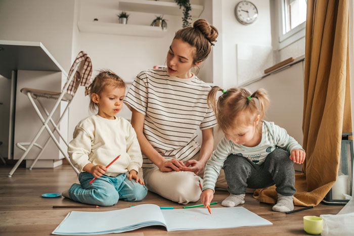Teen babysitter in a striped shirt with two kids on the floor, coloring together in a cozy room.