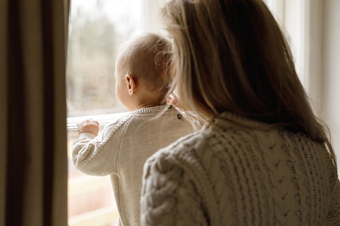 Woman with child at a window, both in knitted sweaters, symbolizing family time and unexpected stays. Woman with child at a window, both in knitted sweaters, symbolizing family time and unexpected stays.