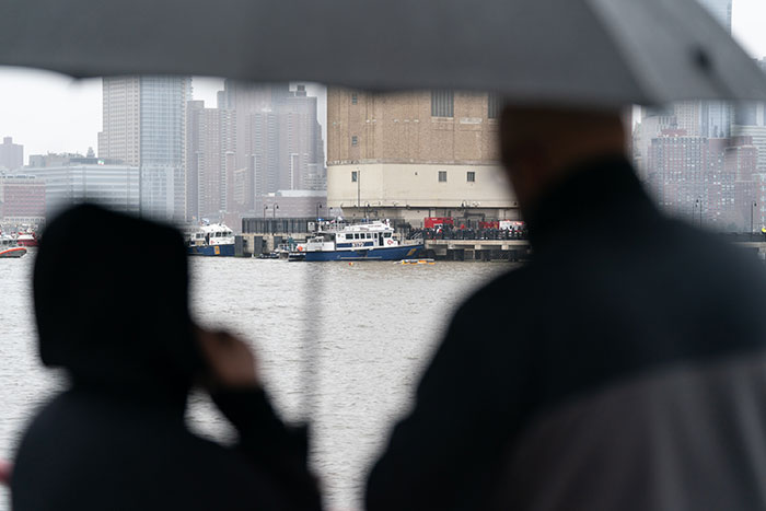 Silhouettes of onlookers near NYC helicopter crash site by the river under an umbrella, with rescue boats in the background. Silhouettes of onlookers near NYC helicopter crash site by the river under an umbrella, with rescue boats in the background.