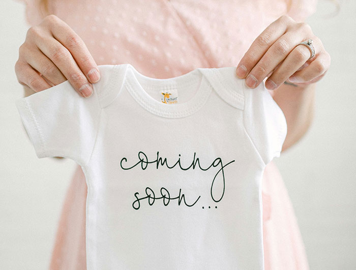 Person holding a baby onesie with "coming soon," relating to name change secrecy. Person holding a baby onesie with "coming soon," relating to name change secrecy.