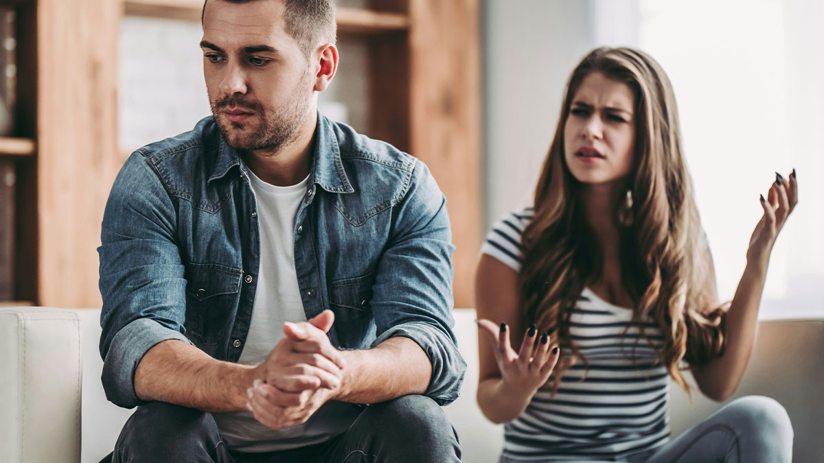 Man giving girlfriend silent treatment while she gestures in frustration during tense conversation in living room.