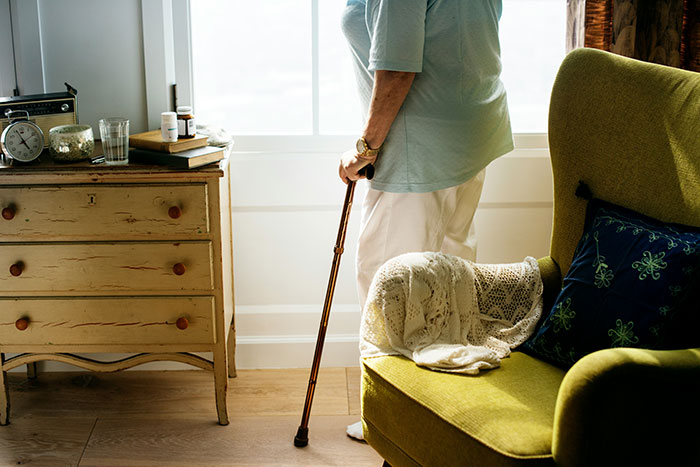 Elderly person with cane beside a chair and dresser in brightly lit room. Elderly person with cane beside a chair and dresser in brightly lit room.
