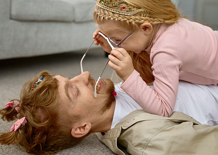 Father playfully lying on the floor with his child, who is wearing a princess tiara and glasses, showcasing family time. Father playfully lying on the floor with his child, who is wearing a princess tiara and glasses, showcasing family time.