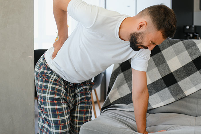 Man in pain holding his back, wearing white t-shirt and plaid pants, near a sofa. Man in pain holding his back, wearing white t-shirt and plaid pants, near a sofa.