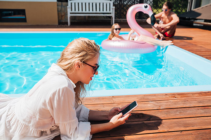 Woman in sunglasses using phone by pool while neighbors’ relatives enjoy pool with inflatable flamingo nearby. Woman in sunglasses using phone by pool while neighbors’ relatives enjoy pool with inflatable flamingo nearby.