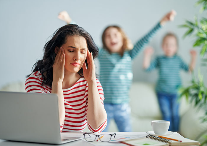 Stressed woman holding her head with kids playing wildly behind her, relating to charging friend for furniture damage. Stressed woman holding her head with kids playing wildly behind her, relating to charging friend for furniture damage.