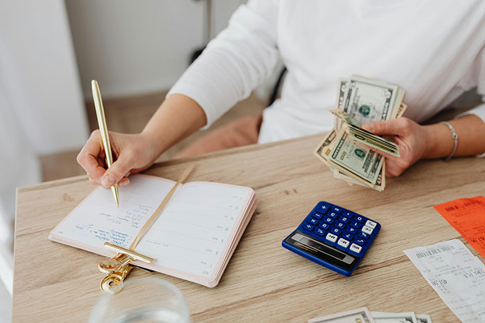 Person counting money and writing in planner with calculator and receipts on wooden table, spouse secret concept