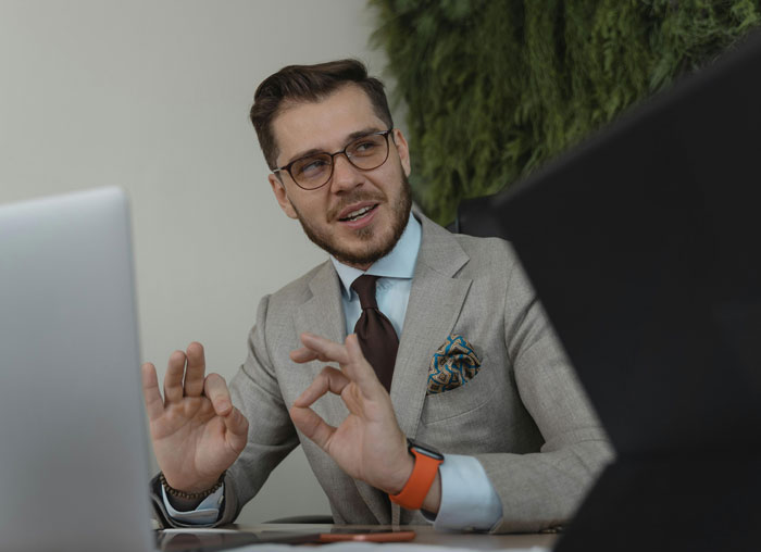 Man in a gray suit and glasses gesturing while discussing infuriating things said by people in charge during a meeting.