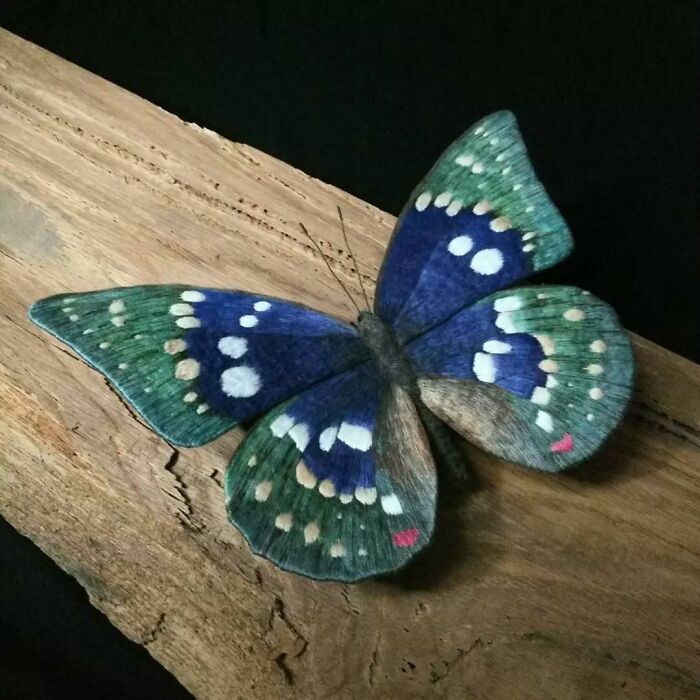 Embroidered fiber butterfly with blue, green, and white patterns resting on a wooden surface, showcasing detailed textile art.