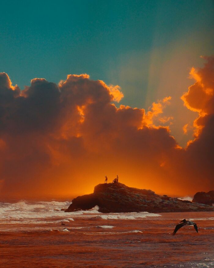 Surreal photo manipulation of people on rocky shore with glowing orange clouds and ocean waves at sunset.