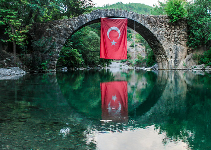 Stone arch bridge over clear water with Turkish flag hanging, reflecting on the river in a lush green landscape about living in home countries.