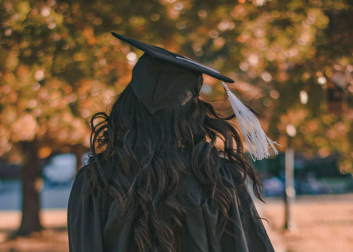 Graduate with long hair wearing a black cap and gown walking outdoors, representing student loan collections impact. Graduate with long hair wearing a black cap and gown walking outdoors, representing student loan collections impact.