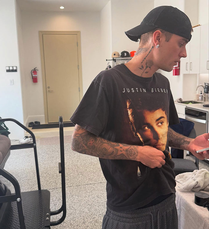 Justin Bieber wearing a black cap and graphic t-shirt indoors, checking his phone, showing tattoos on his arms and neck. Justin Bieber wearing a black cap and graphic t-shirt indoors, checking his phone, showing tattoos on his arms and neck.