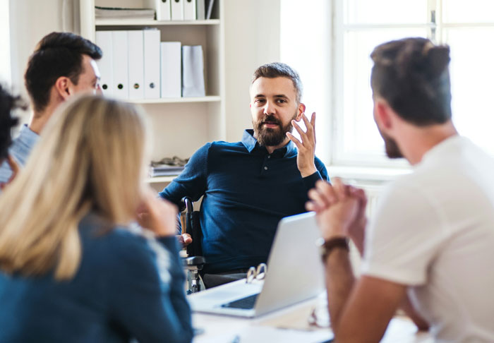 A group of people in a meeting room listening to a man in charge discussing ideas around a laptop.