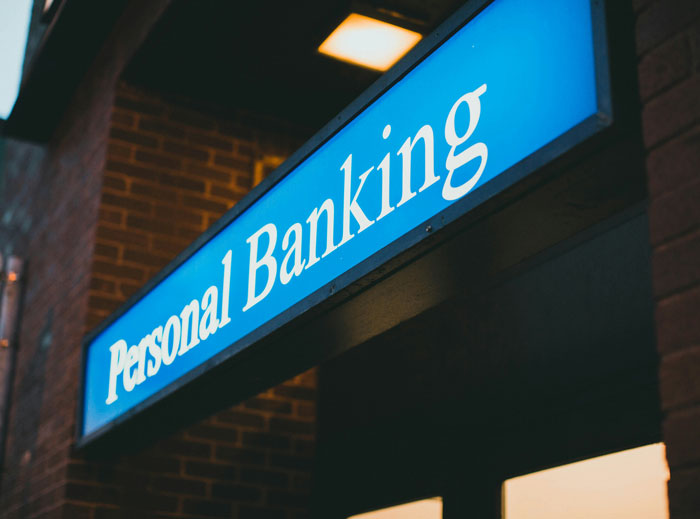 Blue illuminated personal banking sign on a brick building highlighting saving money strategies that cost more.