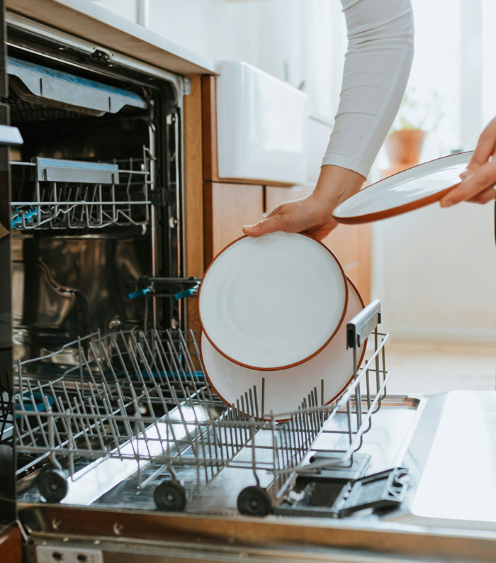 Person loading clean plates into a dishwasher illustrating things people do to save money that cost them more.
