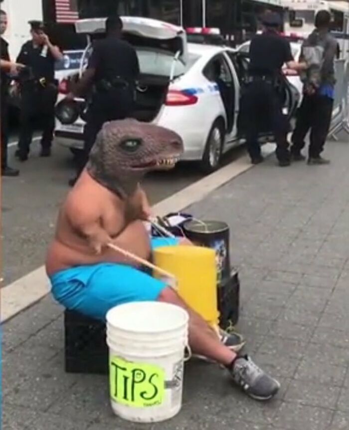 Person with a dinosaur mask playing drums on street with a tips bucket nearby and police officers in the background cursed images