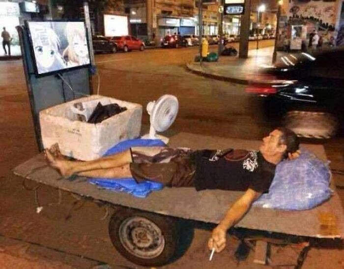 Man lying on makeshift bed on street cart watching TV with fan nearby in a cursed image for a weird brain break.